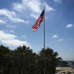 American flag waves atop a flagpole above palm trees and buildings, with water and a cloudy blue sky in the background.