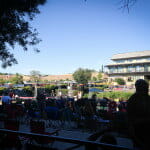 Crowd at an outdoor 4th of July event, with people seated and mingling under trees, a building and open fields in the background.