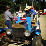 Vintage blue 1913 Model T Ford with the number 123 on its front, parked outdoors. A man is riding, a girl is sitting next to him, and another man is standing next to her.