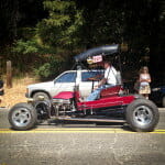 Man driving a 1923 T-Bucket Roadster Blown Ford with an open engine and black canopy roof on a sunny road, passing parked cars and pedestrians.