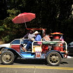 Vintage blue 1913 Model T Ford, decorated with colorful objects and an umbrella, with a man riding, a girl sitting next to him, and another man standing next to her, along a sunny street with trees in the background.