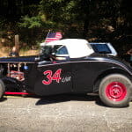 1934 Ford Classic Hot Rod Roadster with red wheels and number 34, parked on a road. A man sits in the car holding the steering wheel, with two women and a girl under trees in the background.